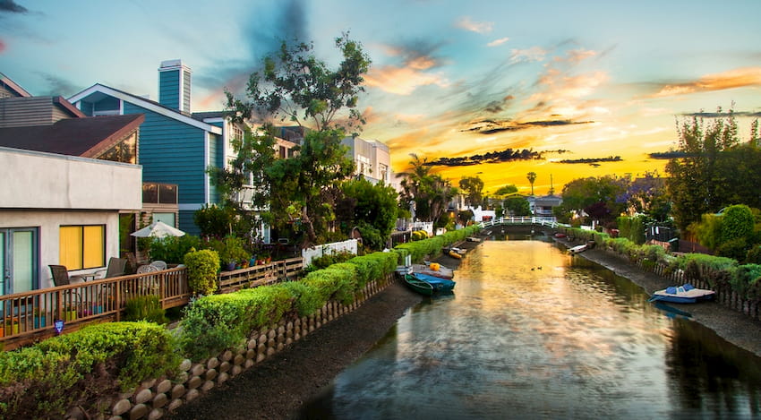 the sun sets over a canal in Venice, California. The bands are lined with kayaks, and a bridge is visible in the background