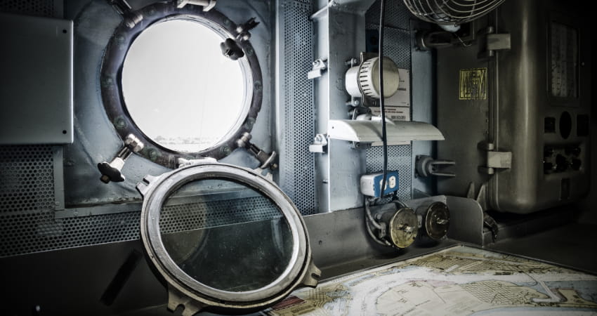 Interior of a mid-century ship, with an open porthole, pipes lining the walls, and mechanical instruments strewn across a desk