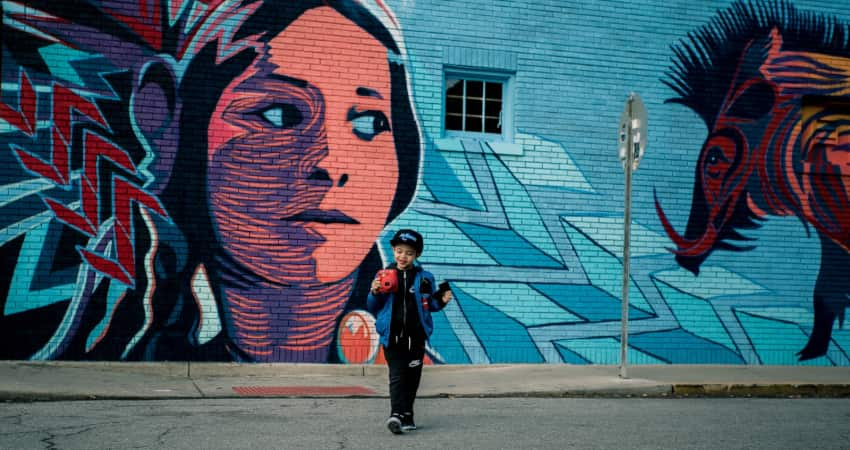 A child holds a camera and stands in front of a bright mural. The mural depicts a Native American woman and a boar on a blue background