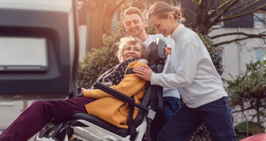 Two people assisting a elderly person in a wheelchair