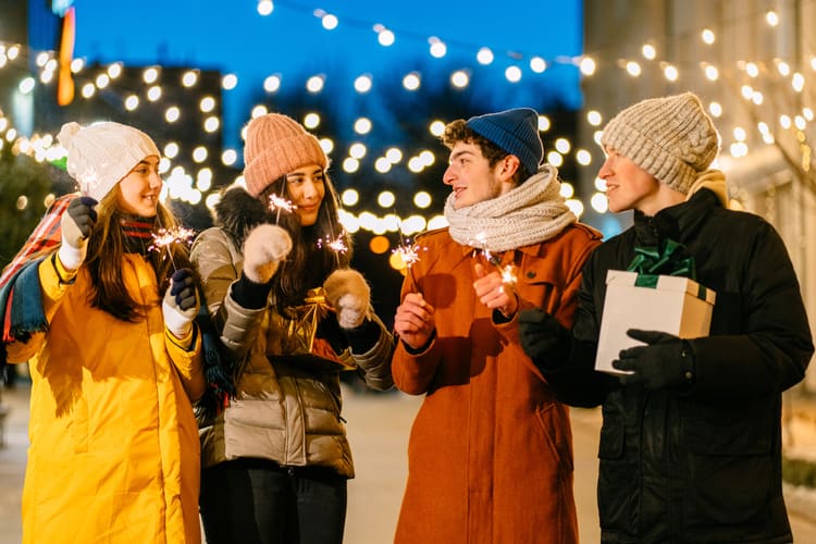 four friends hold sparklers while wearing coats and hats with snow and holiday lights in the background