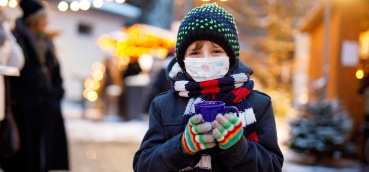 Child wearing hat, gloves, and COVID-19 protective mask while holding hot chocolate at a Christmas market