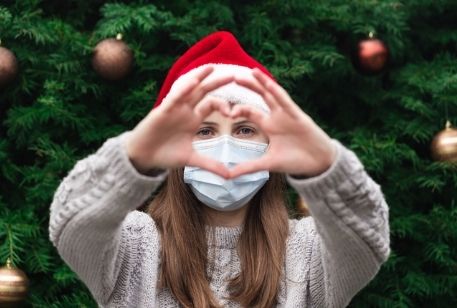 Girl wearing a Santa hat and COVID-19 face mask standing in front of a Christmas tree and making a heart with her hands