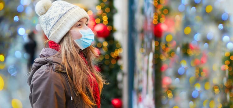 a woman with a beanie hat and facemask looks at a holiday store display in a window