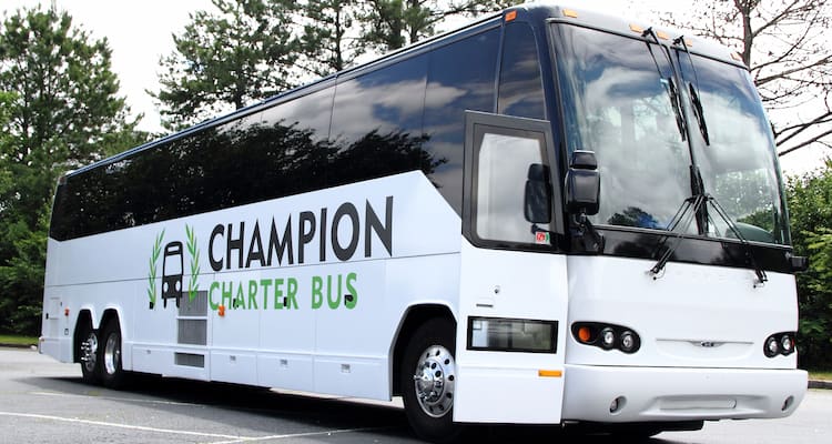 A white branded charter bus with a Champion Charter Bus logo parks in a wooded lot